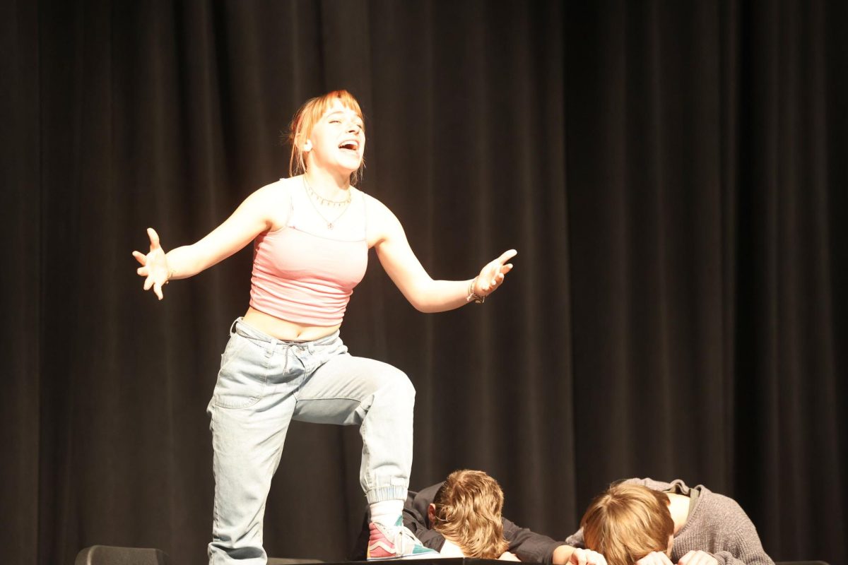 Senior Skye Rodman cheerfully performs on top of the table while juniors Kevin Zehms and Grant Tucker are sleep in their ensemble acting "Sports"  Feb.15.